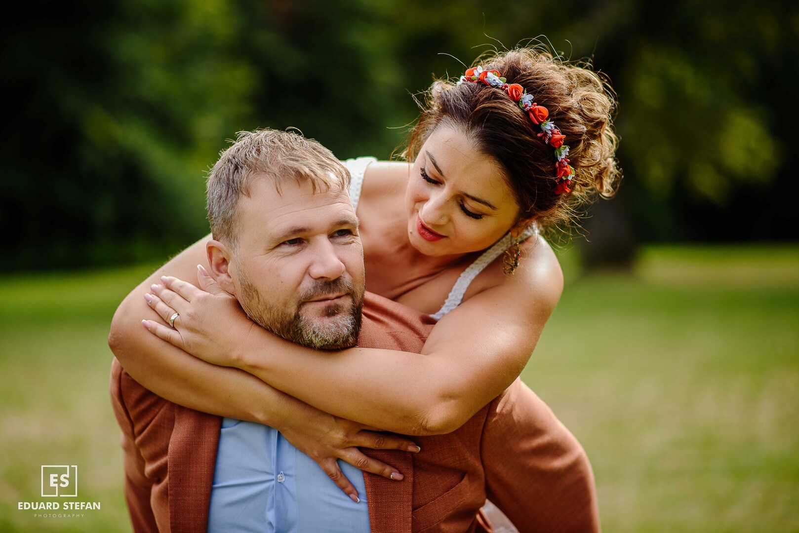Woman with a floral headband playfully hugging her partner from behind in a sunny park setting.